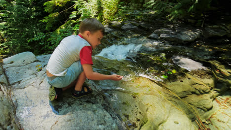 young boy kid crouched down next to waterfall on top of rocky cliff in nature park, british columbia canada