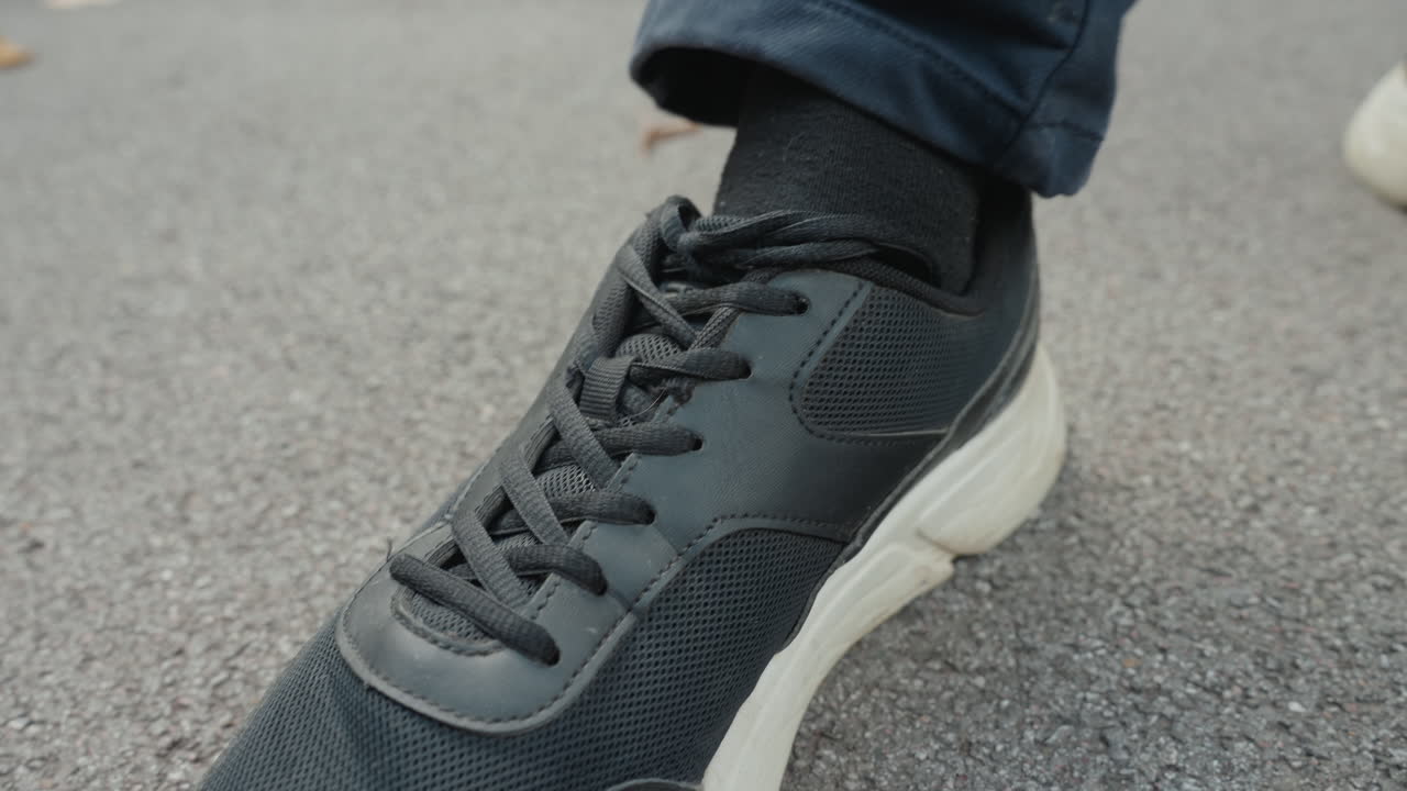 Primo piano di scarpa da ginnastica da uomo su marciapiede con laccio slacciato svolazzante, texture di asfalto urbano, jeans risvoltati, esitazione impercettibile di un pendolare in pausa, runner che controlla il passo, studente che modifica la postura, addetto alle consegne