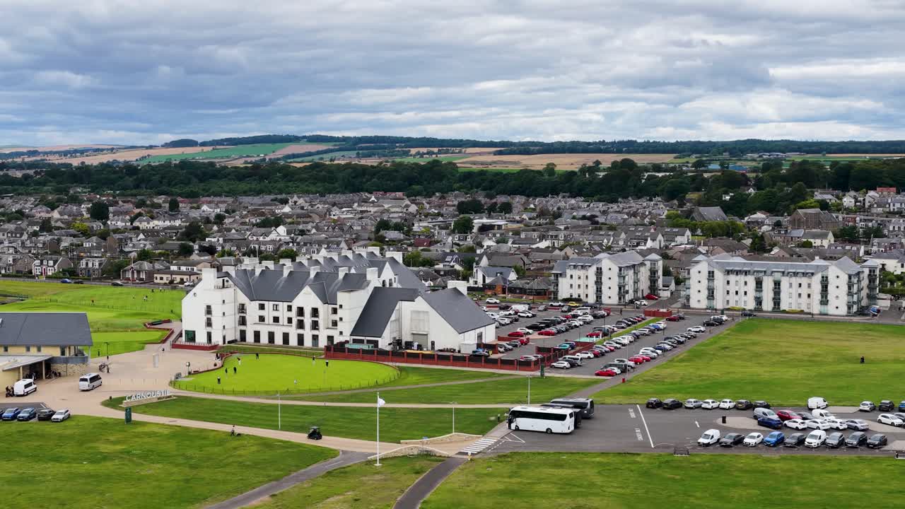 Drone camera glides above golf clubhouse, fairway, parking lot, and surrounding town under cloudy skies