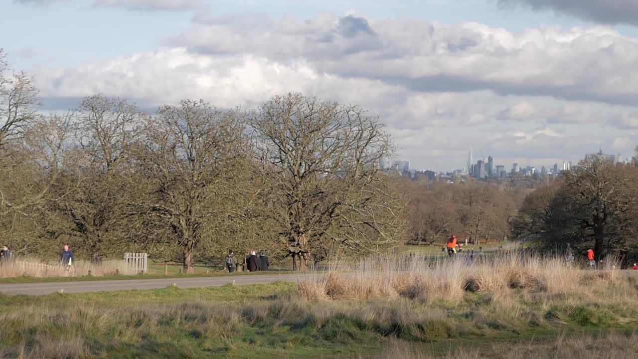Clear autumn day in Richmond park, London, while people jog, walk and cycle. City skyscrappers on the horizon. Big white clouds on sky.