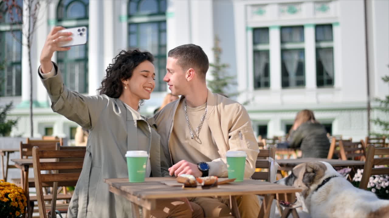 Una coppia felice all'aperto vicino a un bar. Selfie, baci, caffè sul tavolo, il loro cane nelle vicinanze. Atmosfera autunnale