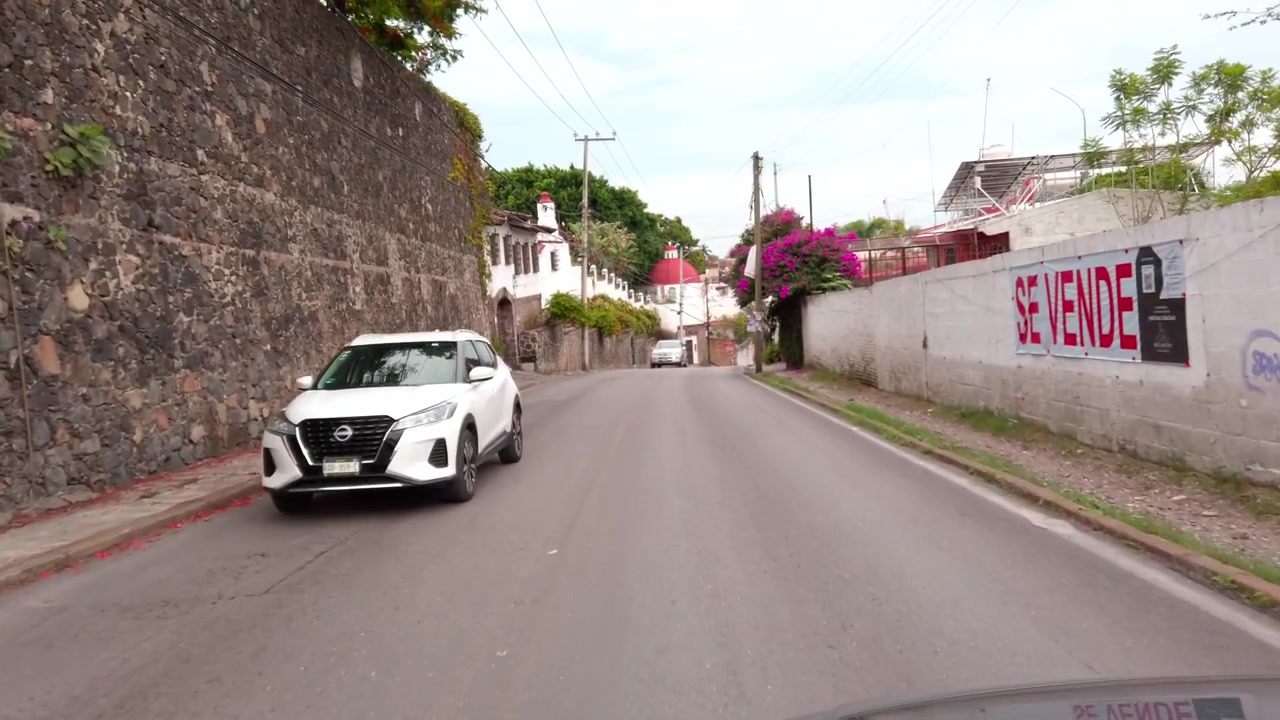 Vista dall'auto che guida lungo una strada residenziale con muri di confine e pali di servizio. La strada asfaltata costeggia case del quartiere, ingressi recintati e vegetazione a bordo strada sotto linee elettriche aeree.