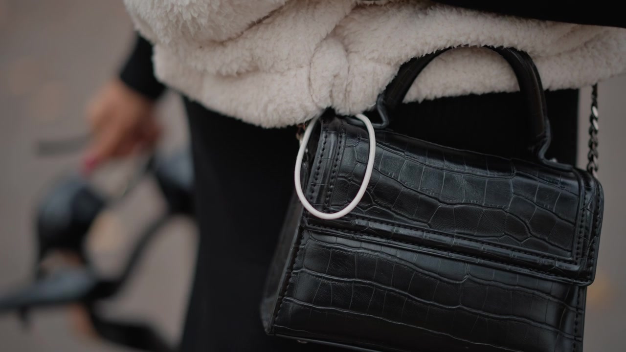 Borsa in pelle nera con stampa coccodrillo che oscilla all'altezza dell'anca durante il movimento di camminata su una strada urbana, con tracolla a catena, anello in metallo, cerniera, cuciture e uno stile di moda testurizzato su uno sfondo morbido e sfocato.