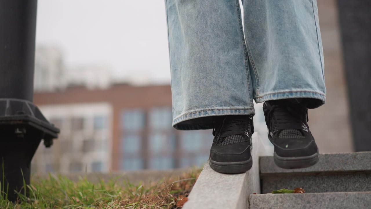 Primo piano dei piedi di una persona con jeans azzurri e scarpe da ginnastica nere che scendono con cautela gradini di pietra con erba visibile in basso. Lo sfondo presenta edifici urbani sfocati che indicano