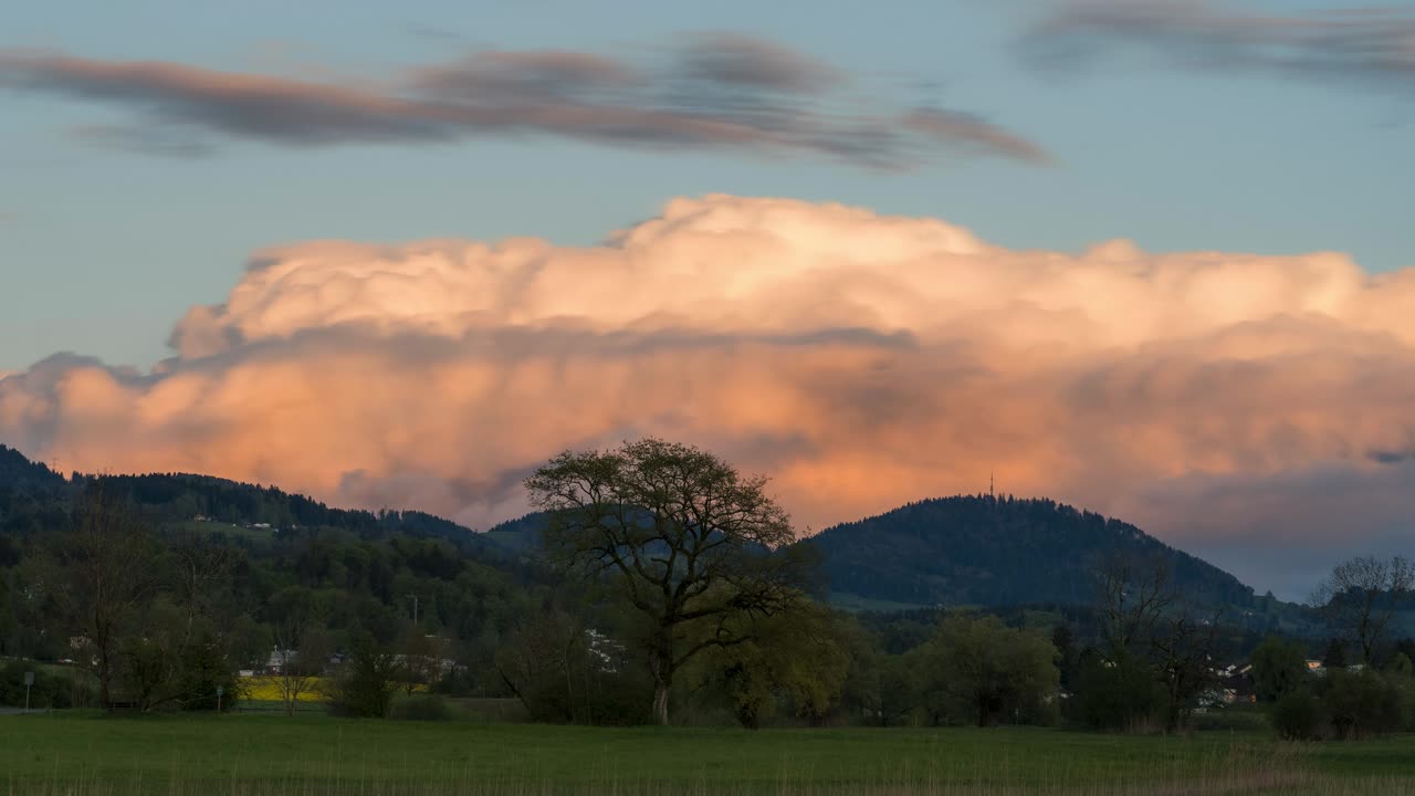 Clouds swirl and drift across the natural landscape at golden hour. The clouds are illuminated by yellow and pink. It's spring, and everything is blooming. Mountains can be seen in the background.