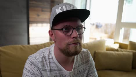 millennial man with beard watching television fixes his baseball cap, zoom in