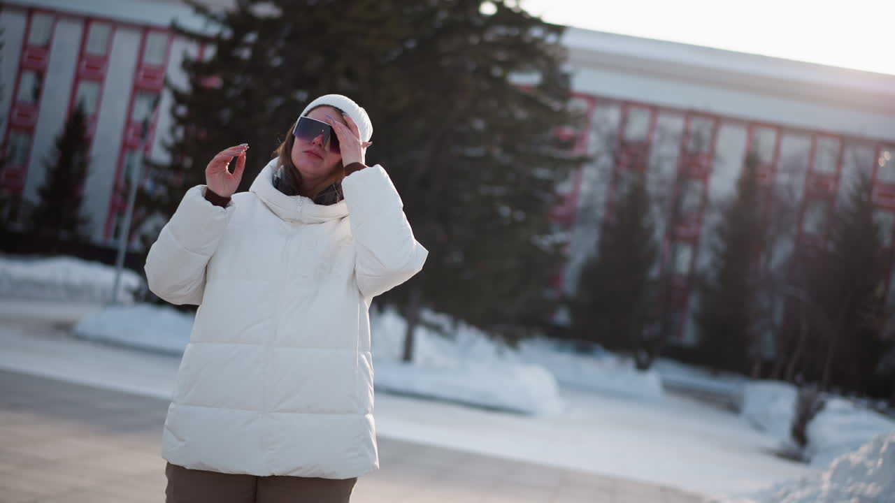 Young woman stands in snowy park under bright sun wearing white beanie and puffer coat, adjusting tinted black sunglasses, surrounded by bare trees and soft winter light, cheeks rosy from cold breeze