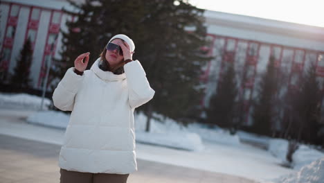young woman stands in snowy park under bright sun wearing white beanie and puffer coat, adjusting tinted black sunglasses, surrounded by bare trees and soft winter light, cheeks rosy from cold breeze