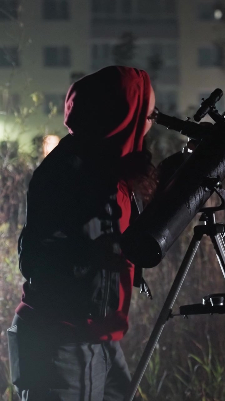 Donna con felpa rossa con cappuccio e giacca nera osserva. La persona con la giacca scura guarda attentamente attraverso la lente di un cannocchiale. Erbe alte ondeggiano dietro di lei, suggerendo un ambiente esterno.