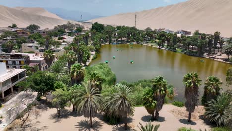 Oasis-Del-Desierto-Huacachina,-Perú-Con-Lago-Y-Palmeras,-Con-Grandes-Dunas-De-Arena-En-El-Fondo