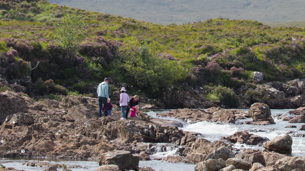 Family walks along rocky riverbank, lush green hills, bright daylight, wide shot, steady camera