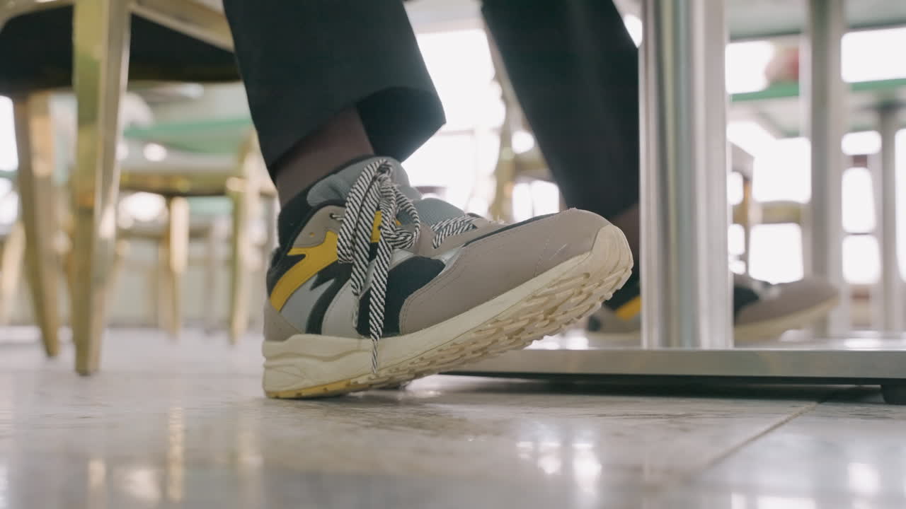 Nærbillede af sneakers under bordet viser en sort kvindelig professionel, der skubber foden ind under bordet under en afslappet frokostdate, bistrostole, flisegulv, skarpt middagslys, selvsikker legende forbindelse.