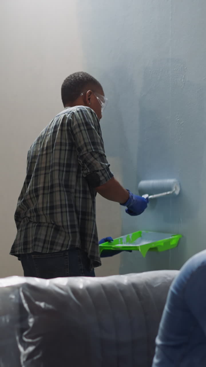 Black man in checkered shirt applies paint on wall in living room. African-American friend fixes chair sitting on sofa. Reconstruction of apartment