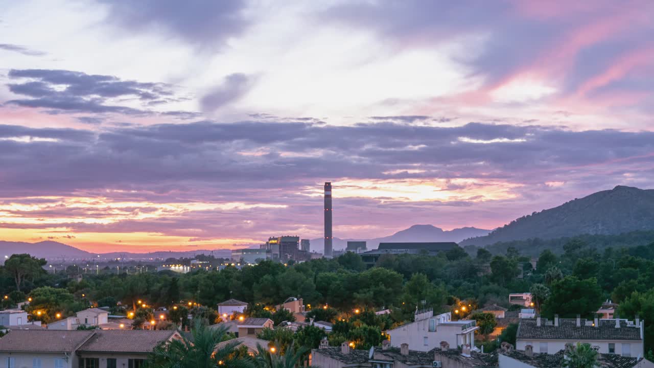 Timelapse of a dramatic sunset over a town, featuring an industrial smokestack and mountains