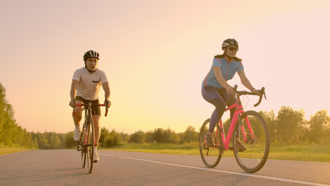 Handsome bearded professional male cyclist riding his racing bicycle in the morning together with his girlfriend both wearing protective helmets and eyeglasses sun shining through between them
