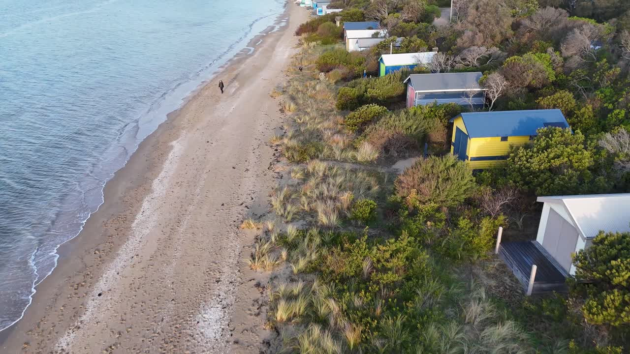 Drone glides above sandy beach, vibrant bathing boxes, coastal vegetation, and calm sea at sunset