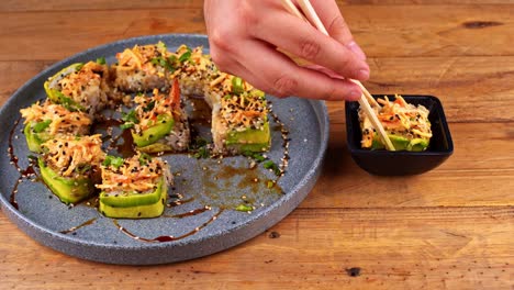 close-up of hands with chopsticks, taking an avocado sushi roll on a blue plate and dipping it into eel sauce, but the roll sinks in the sauce