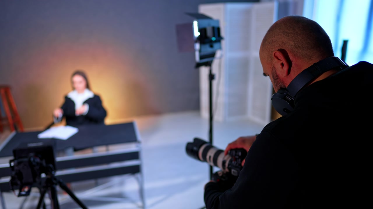 Fotograf tager billeder af model under studievideooptagelse. Over skulderen-udsigt af fotograf, der holder kamera fokuseret på kvinde, der sidder ved et skrivebord