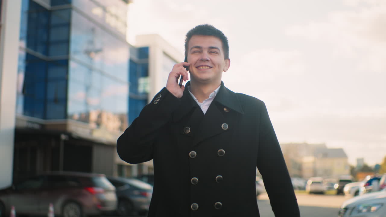 Investor in black coat talking on phone with excitement and pointing in urban area with modern glass buildings, soft sunlight, parked cars, and lively outdoor setting