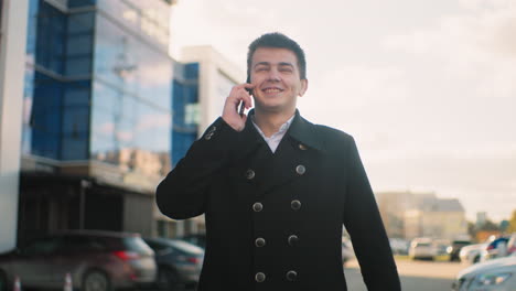 inversor en abrigo negro hablando por teléfono con emoción y señalando en una zona urbana con edificios modernos de vidrio, luz solar suave, coches estacionados, y un entorno al aire libre animado