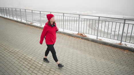 woman in red winter jacket walking along interlocked pathway with bag over shoulder and focused expression near iron railing, foggy atmosphere, and distant car below bridge in background