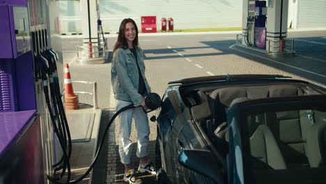 A-beautiful-young-brunette-girl-in-a-denim-jacket-refuels-her-dark-gray-convertible,-she-smiles-and-looks-at-the-camera