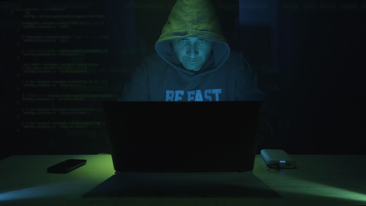 Male Hacker In Hood Working On Laptop In A Dark Room At Night. - zoom in