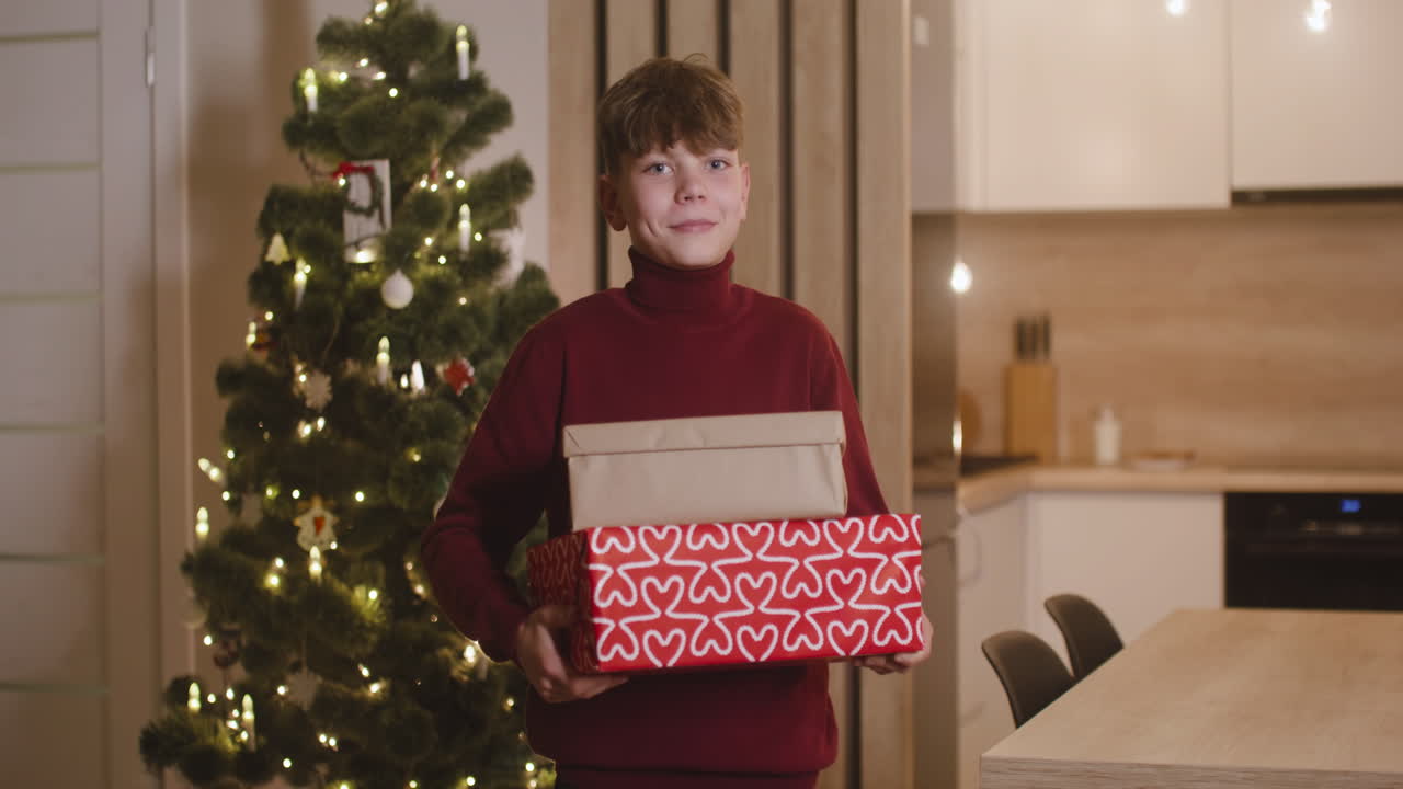 vista frontal de un niño rubio con suéter de cuello alto rojo sosteniendo regalos en una habitación decorada con un árbol de navidad