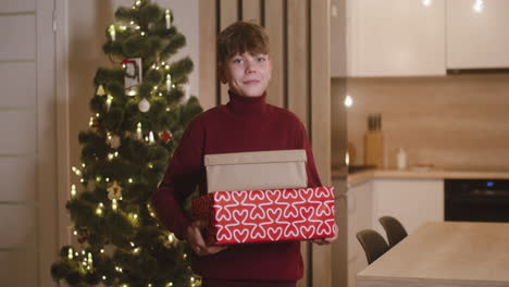 front view of a blond boy in red turtleneck sweater holding gifts in a room decorated with a christmas tree