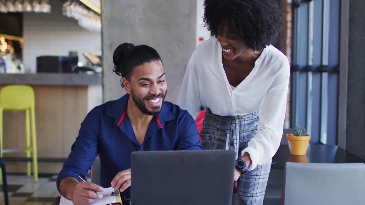 Diverse couple sitting at a table in a cafe using laptop smiling and talking