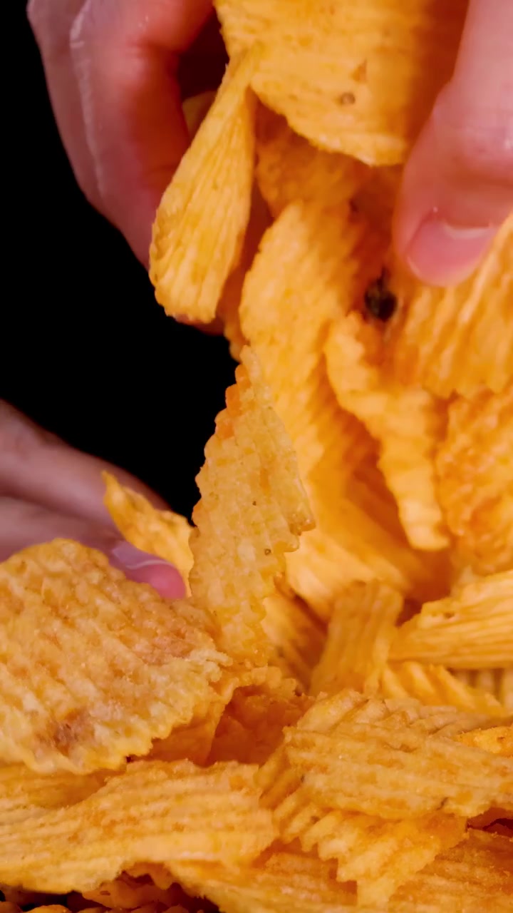 Close-up de uma mão pegando e soltando batatas fritas onduladas, iluminação dramática, fundo preto.