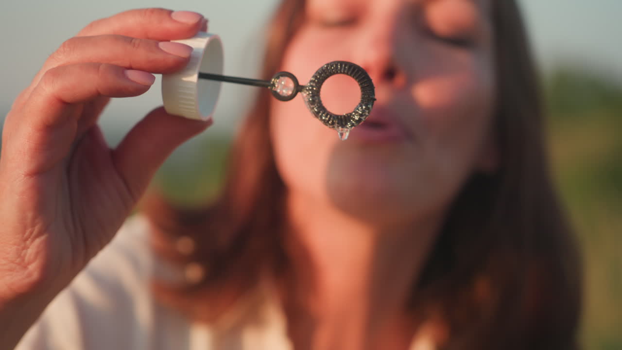 Close up of fair skin hand holding bubble wand dipped in soapy solution with rainbow sheen, soft evening light glinting off ring and highlighting delicate detail in tranquil outdoor setting