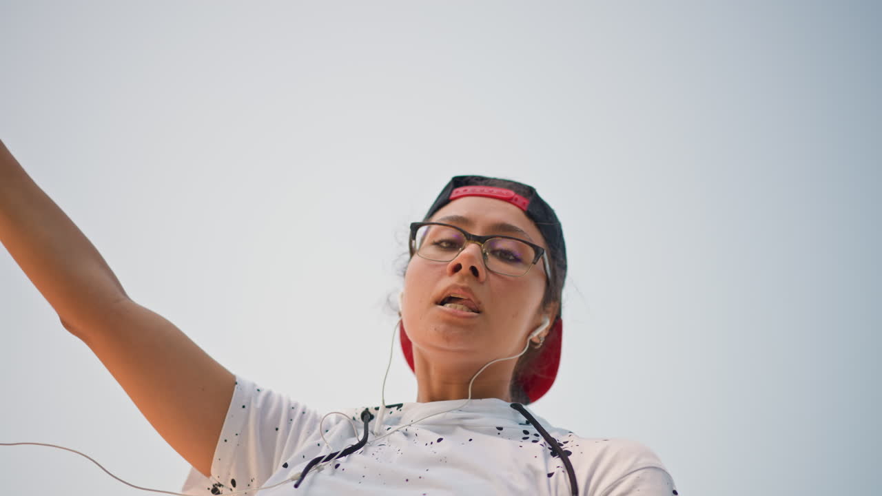 Celebración en un parque desde ángulo bajo con un teléfono y los brazos extendidos; un joven con gorra y auriculares captura un movimiento alegre con el cielo abierto de fondo; perspectiva cinematográfica con gestos dramáticos y ambiente enérgico, gratis.