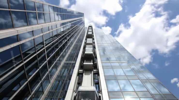 Un punto de vista dinámico de un edificio moderno de gran altura con fachada de cristal reflectante y ascensores verticales, capturado desde abajo bajo un cielo nublado.