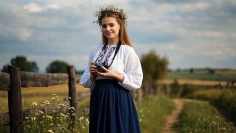 young woman in traditional attire with flower crown holding a camera in a rural landscape