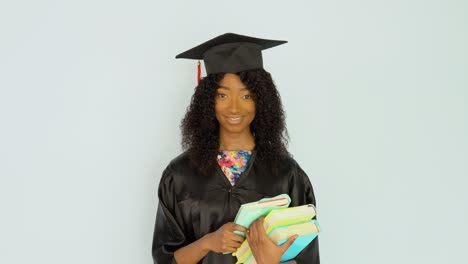 una joven afroamericana con una túnica negra y un sombrero de maestro se mantiene erguida sosteniendo libros de texto. mira a la cámara. retrato de media altura