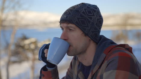 a man in a snow hat calmly sips tea out of a mug with a snowy backdrop, slow motion