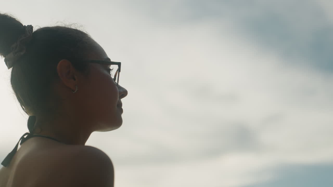 Perfil de jovem mulher contra céu nublado, usando óculos e coque, contraluz cinematográfica de baixo ângulo, brilho dourado suave, roupa de praia, brisa suave, expressão introspectiva, clima de noite de verão