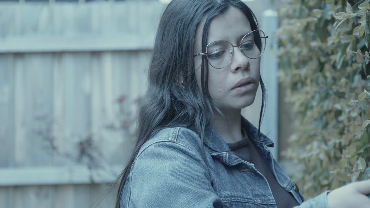 A young woman with glasses tends to garden in a tranquil setting. The overcast lighting casts a serene mood as she interacts with greenery. Denim jacket adds a casual vibe