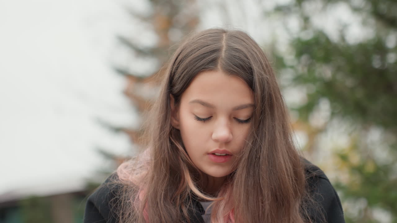 white teenage student outdoors studying notes, closeup profile shots of white girl with long brown hair and pink streaks, black coat and chilly overcast sky, autumn trees and scattered leaves
