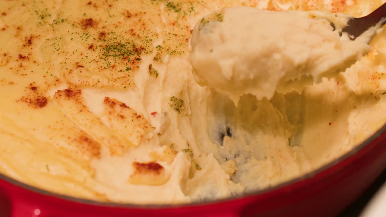Un primo piano mostra un cucchiaio che preleva purè di patate cremoso da una pentola rossa.