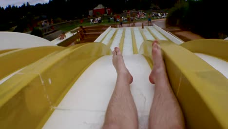 young man sliding down a yellow water slide in amusement par, public bath and landing in a splash of water, unrecognizable, legs only