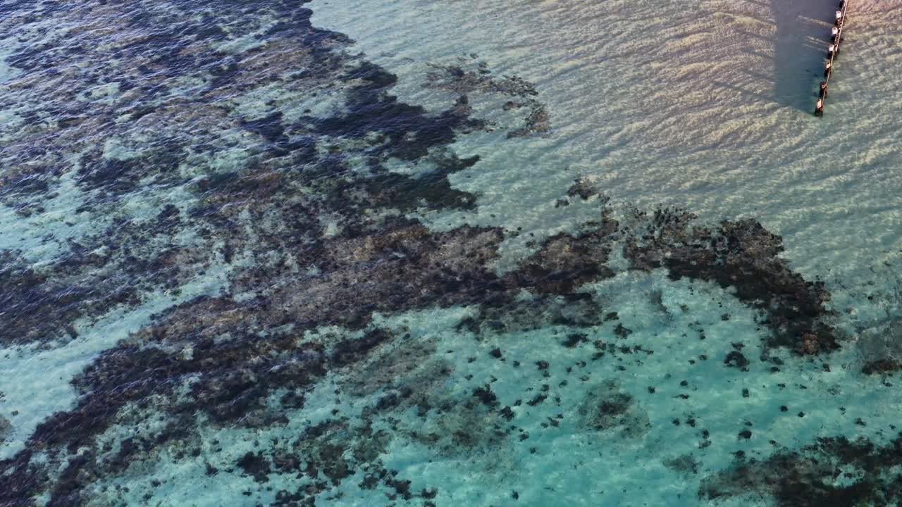 Drone glides above sandy shoreline, pier, and clear water revealing underwater reefs at golden hour