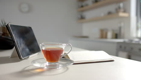 a tablet and cup of tea rest on a kitchen counter beside open notebook