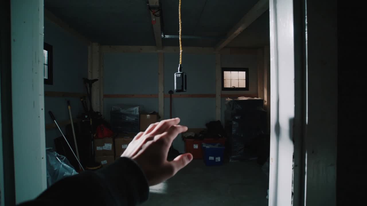 Entrando attraverso una porta, una mano destra con una manica scura si allunga, tirando un cordino giallo per accendere la luce del garage.