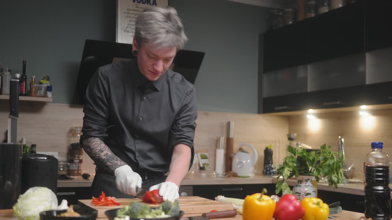 joven chef profesional con una elegante camisa negra con un aspecto alternativo, tatuajes, guantes rebanando pimientos rojos una cocina moderna y elegante con verduras frescas además de deslizamiento cinematográfico 4k