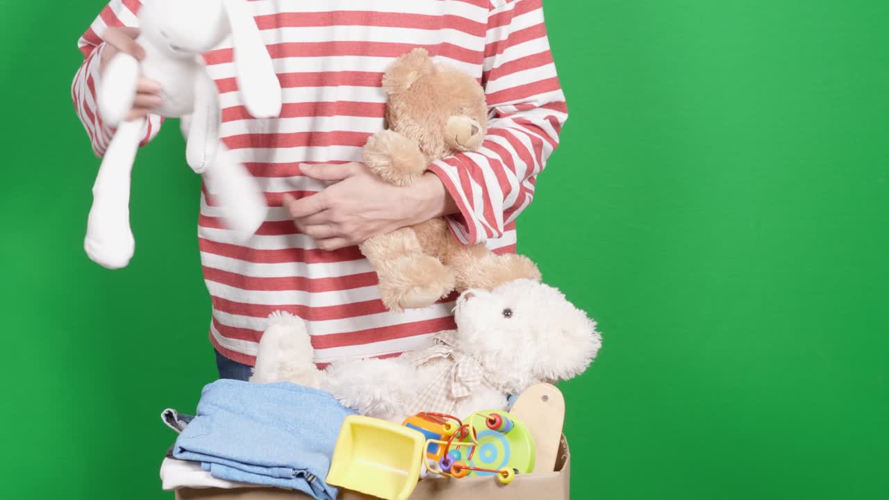 자선 기부를 위해 옷과 장난감을 준비하는 여성. 여성 손이 어린이 장난감을 분류하고 포장하고, 어린이 옷을 녹색 화면 위의 카드보드 상자에 넣습니다.