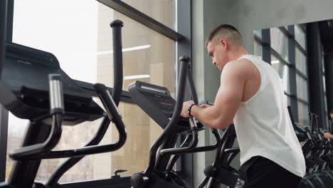 Side view of man gripping elliptical trainer handles and checking console, intense focus and sweat in modern gym studio with glass windows reflecting exercise scene and athletic posture concentration