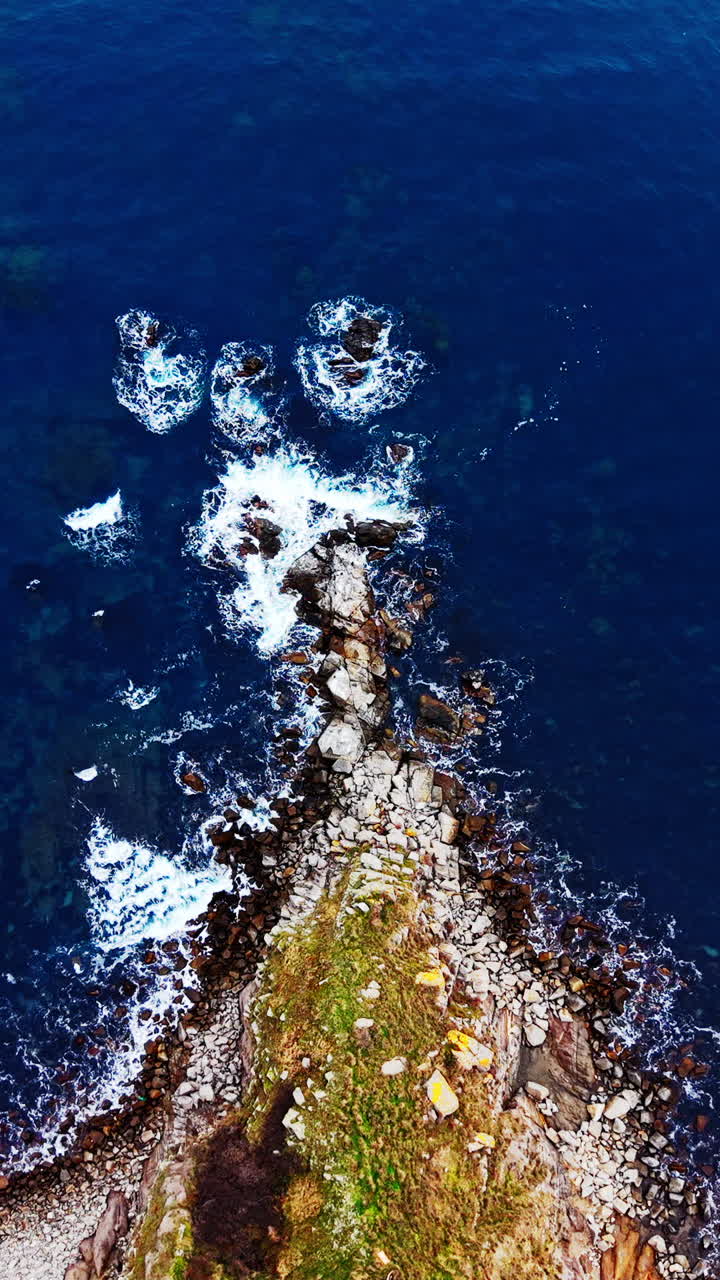 Sharp stone cliff covered with grass surrounded by deep blue ocean water. Aerial view of rocky peninsula jutting into blue sea. Vertical video