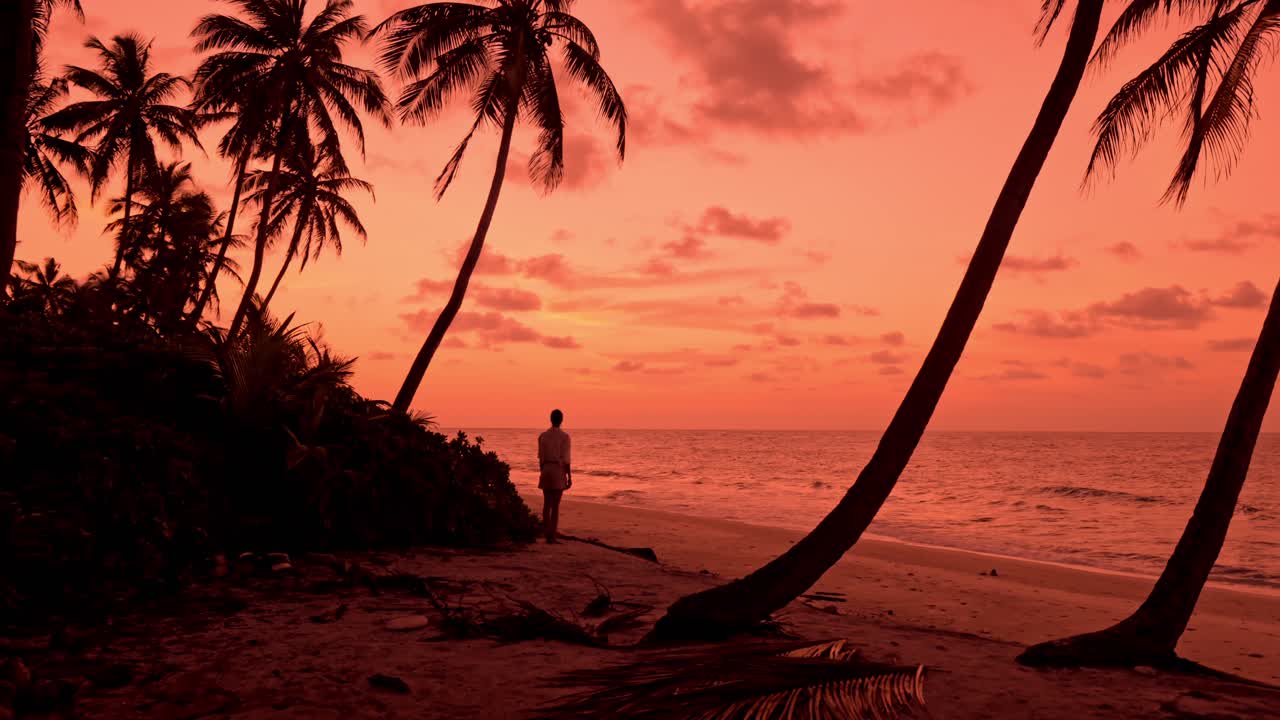 A breathtaking view captures a lone woman strolling along a pristine, palm-lined beach in Fuvahmulah, Maldives at sunset.